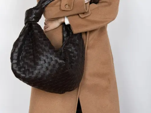 Dunkle, gewebte Tasche mit Knoten-Detail, getragen zu einem Kamelhaarmantel. Sieht aus wie eine Bottega Veneta, aber es ist ein Bottega Veneta Fake.