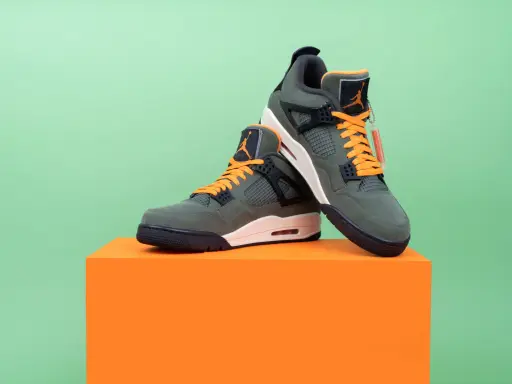 Jordan Motorsport Sneaker in Olivgrün mit orangefarbenen Schnürsenkeln, auf einem orangefarbenen Podest vor hellgrünem Hintergrund.