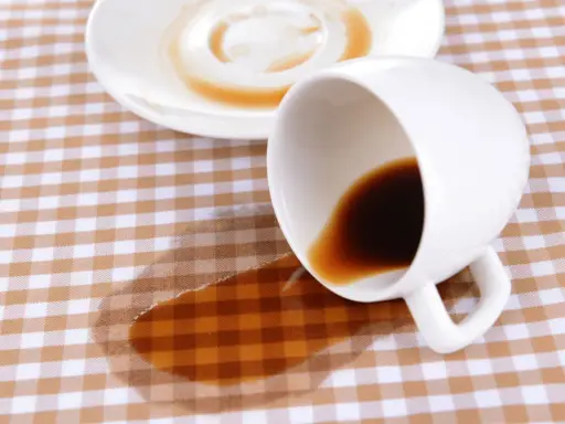 Um Kaffeeflecken zu entfernen, ist hier ein umgekippter Becher mit Kaffee auf kariertem Stoff.