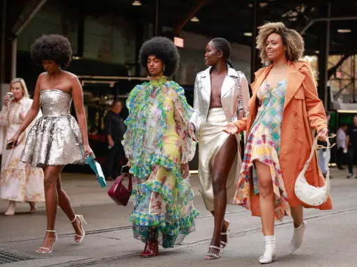 Vier Frauen in auffälligen Outfits. Eine trägt eine silberne Paillettenjacke zum Kleid, eine andere ein buntes Rüschenkleid.