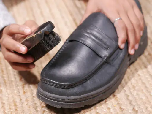 Hände reinigen einen schwarzen Lederschuh mit einer Bürste.