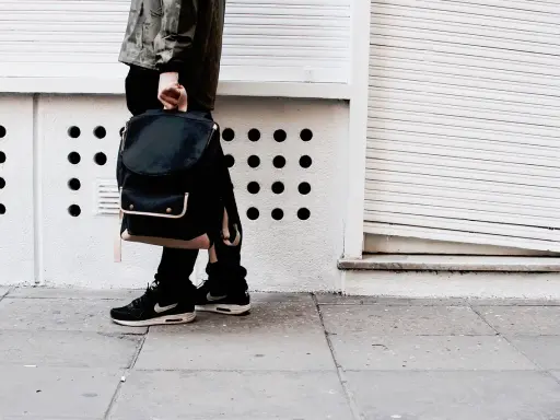 Person mit schwarzem Rucksack und **air max fake** Schuhen steht auf einem Gehweg vor einer weißen Wand mit runden Öffnungen.