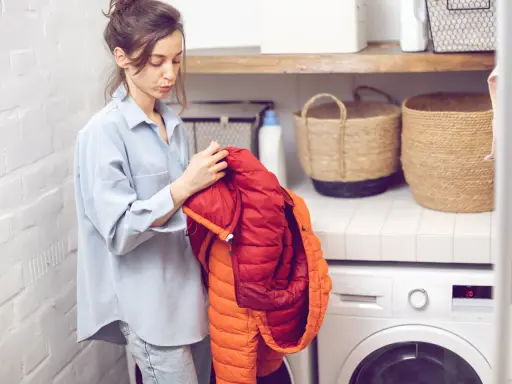 Frau prüft einen roten und orangen Daunenmantel, bereit zum wollmantel waschen. Daneben stehen Wäschekörbe und eine Waschmaschine.