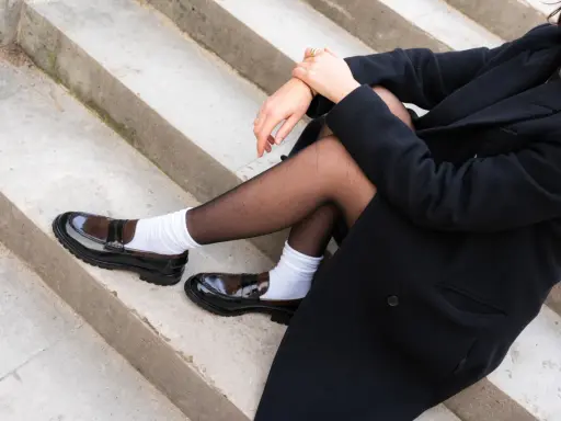 Schwarzer Mantel, schwarze Strumpfhose und weiße Socken mit glänzenden braunen Loafern.