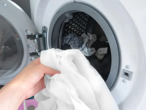 Eine Hand hält ein weißes Hemd, bereit, es in die Waschmaschine zu legen. Die Trommel ist bereits mit anderer Wäsche gefüllt.