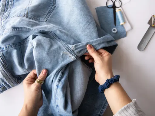 Hände halten hellblaue Jeans, die zum Nähen vorbereitet werden. Nähutensilien wie Schere und Garn liegen bereit. Vielleicht werden die Jeans bald ins Gefrierfach gelegt, um sie zu erfrischen.
