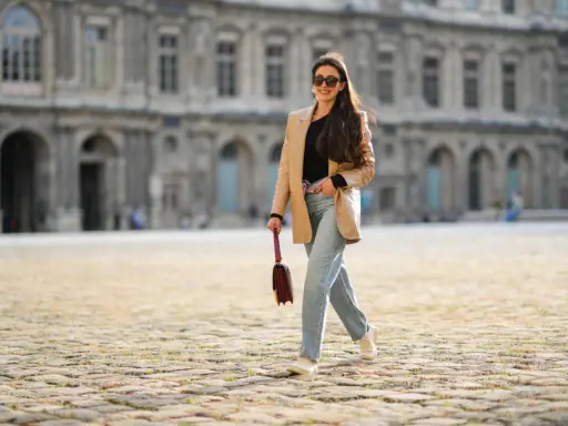 Eine Frau mit langen Haaren und Sonnenbrille spaziert lässig in weiten Jeans und einem beigen Blazer vor einer historischen Fassade.
