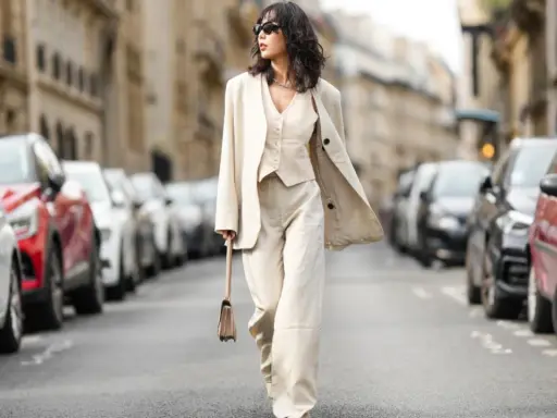 Eine Frau in einem eleganten beigen Hosenanzug mit passendem Westchen und Blazer spaziert durch eine Pariser Straße. Die weiten Hosen und die lässige Silhouette der Schuhe zu Hosenanzug Damen sind perfekt für einen stilvollen Auftritt.