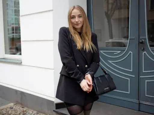 Frau mit blonden Haaren, die ein schwarzes Kleid mit Blazer kombiniert. Sie trägt eine schwarze Tasche und graue Stiefel.