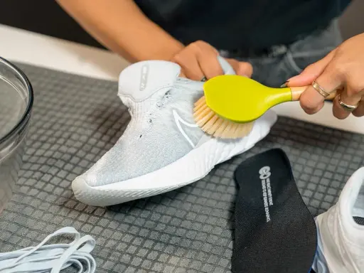 Hände reinigen weiße Mesh-Schuhe mit einer Bürste. Daneben liegen Schnürsenkel und eine Einlegesohle.