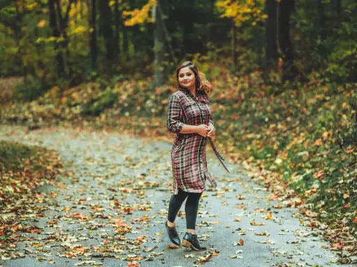 Frau im karierten Kleid mit Leggings steht auf einem herbstlich bedeckten Weg.