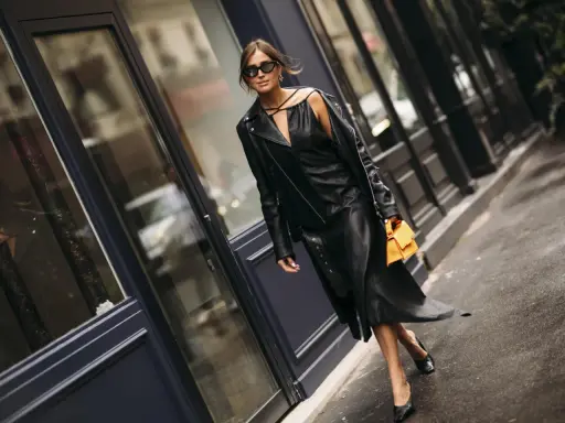 Stilvolle Frau in einem schwarzen Lederkleid und passender Lederjacke, die zeigt, wie man ein schwarzes Kleid kombinieren kann. Orange Handtasche als Farbtupfer.