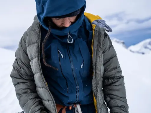 Person in Daunenjacke im Schnee. Die Jacke ist grau, darunter eine blaue Schicht. Perfekt für das Daunenjacke waschen nach der Tour.