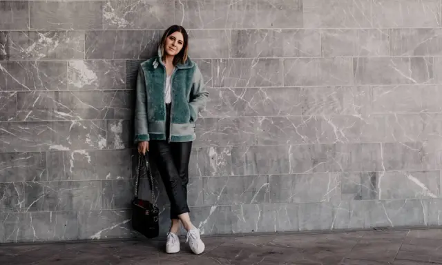 Frau in mintgrüner Lammfelljacke und Lederhose, die weiße Sneakers trägt. Sie steht vor einer grauen Marmorwand.