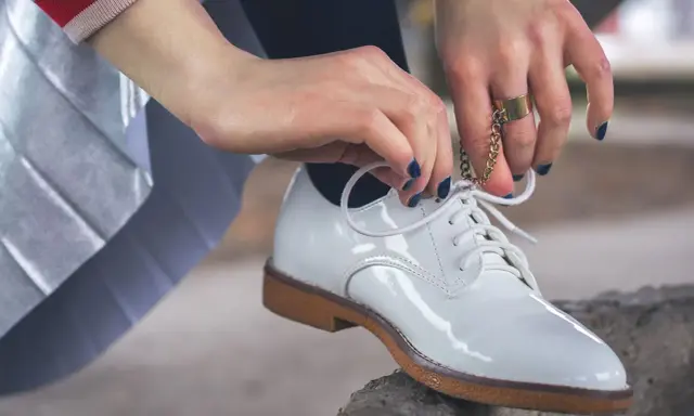 Hände mit dunkelblauem Nagellack binden weiße Lackschuhe. Die Pflege von Lackschuhen ist wichtig, damit sie glänzen.