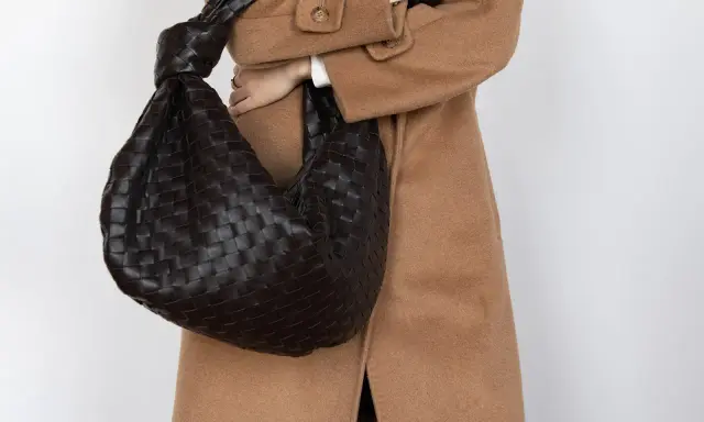 Dunkle, gewebte Tasche mit Knoten-Detail, getragen zu einem Kamelhaarmantel. Sieht aus wie eine Bottega Veneta, aber es ist ein Bottega Veneta Fake.