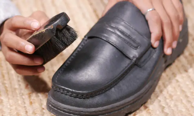Hände reinigen einen schwarzen Lederschuh mit einer Bürste.