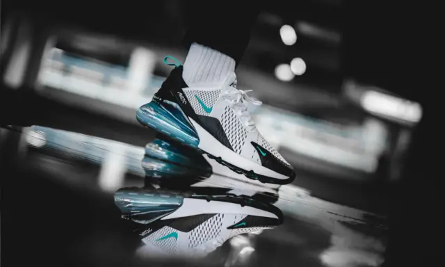 Ein stylischer Nike Air Max 270 in Weiß mit türkisen Akzenten, der auf einer spiegelnden Oberfläche steht. Ein zeitloser Klassiker unter den Nike Air Max alte Modelle.