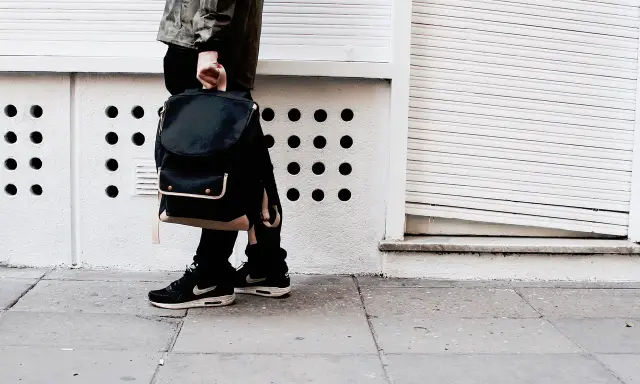 Person mit schwarzem Rucksack und **air max fake** Schuhen steht auf einem Gehweg vor einer weißen Wand mit runden Öffnungen.