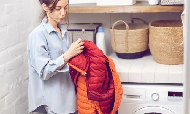Frau prüft einen roten und orangen Daunenmantel, bereit zum wollmantel waschen. Daneben stehen Wäschekörbe und eine Waschmaschine.