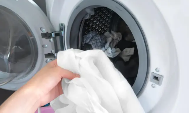 Eine Hand hält ein weißes Hemd, bereit, es in die Waschmaschine zu legen. Die Trommel ist bereits mit anderer Wäsche gefüllt.