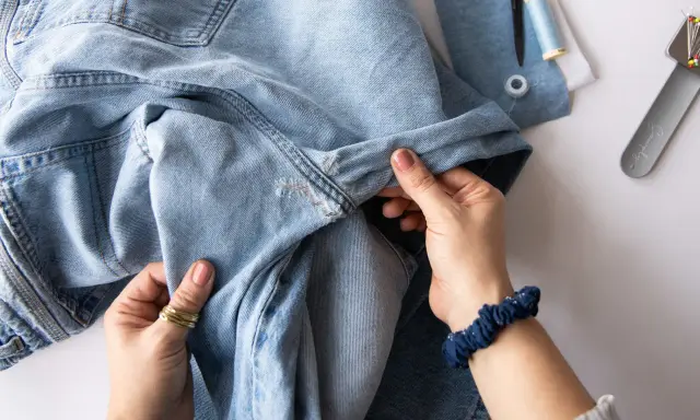 Hände halten hellblaue Jeans, die zum Nähen vorbereitet werden. Nähutensilien wie Schere und Garn liegen bereit. Vielleicht werden die Jeans bald ins Gefrierfach gelegt, um sie zu erfrischen.