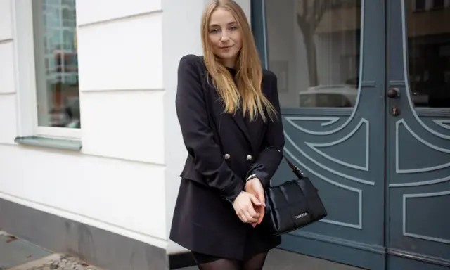 Frau mit blonden Haaren, die ein schwarzes Kleid mit Blazer kombiniert. Sie trägt eine schwarze Tasche und graue Stiefel.