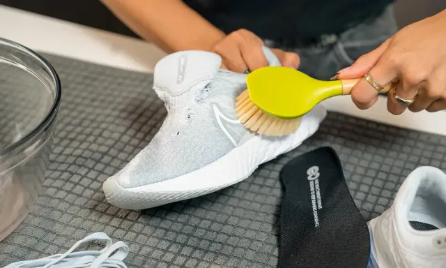 Hände reinigen weiße Mesh-Schuhe mit einer Bürste. Daneben liegen Schnürsenkel und eine Einlegesohle.