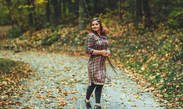 Frau im karierten Kleid mit Leggings steht auf einem herbstlich bedeckten Weg.