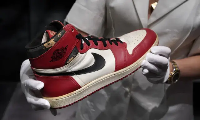 Ein Paar rote und weiße Air Jordan 1 Sneaker, die als die teuersten Schuhe der Welt gelten, werden von Händen in weißen Handschuhen gehalten.