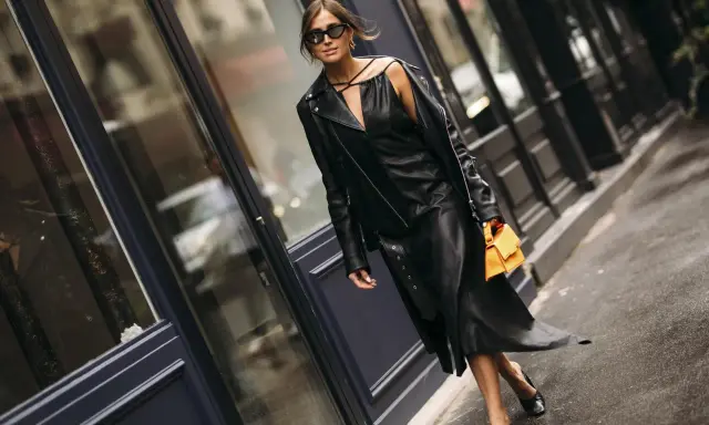 Stilvolle Frau in einem schwarzen Lederkleid und passender Lederjacke, die zeigt, wie man ein schwarzes Kleid kombinieren kann. Orange Handtasche als Farbtupfer.