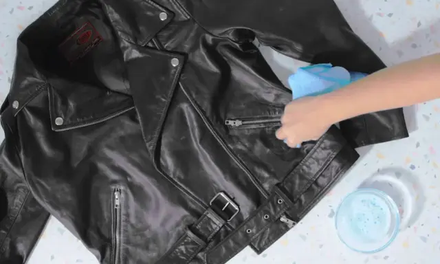 Schwarze Lederjacke reinigen mit einem feuchten Tuch. Eine Hand wischt über die Jacke, daneben eine Schale mit Seifenwasser.