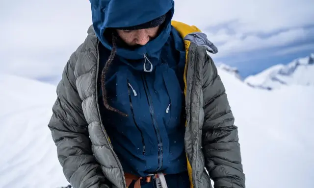 Person in Daunenjacke im Schnee. Die Jacke ist grau, darunter eine blaue Schicht. Perfekt für das Daunenjacke waschen nach der Tour.