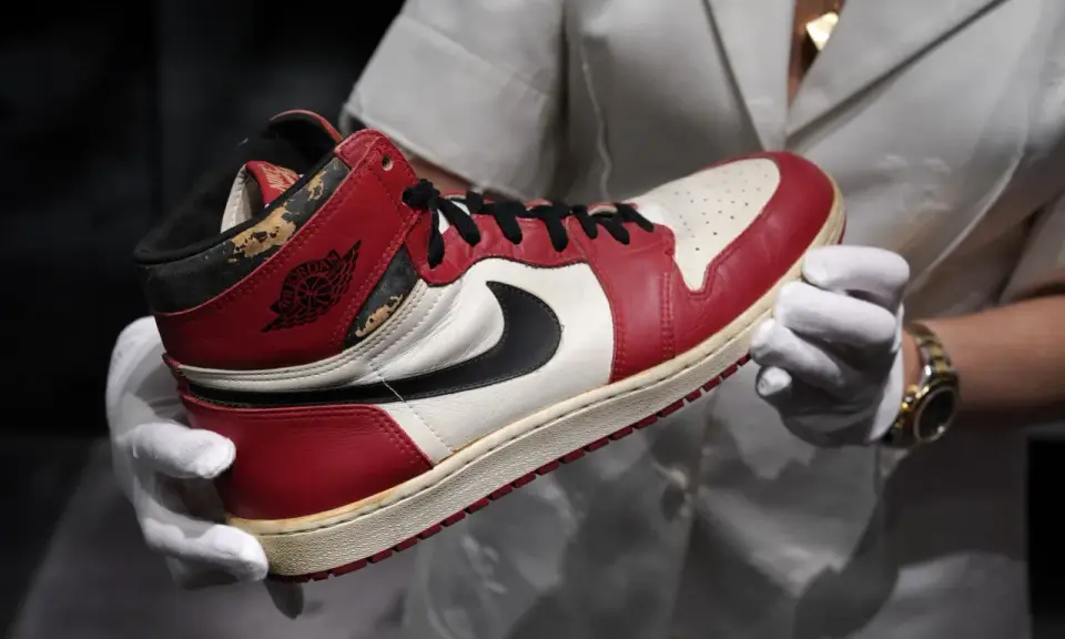Ein Paar rote und weiße Air Jordan 1 Sneaker, die als die teuersten Schuhe der Welt gelten, werden von Händen in weißen Handschuhen gehalten.