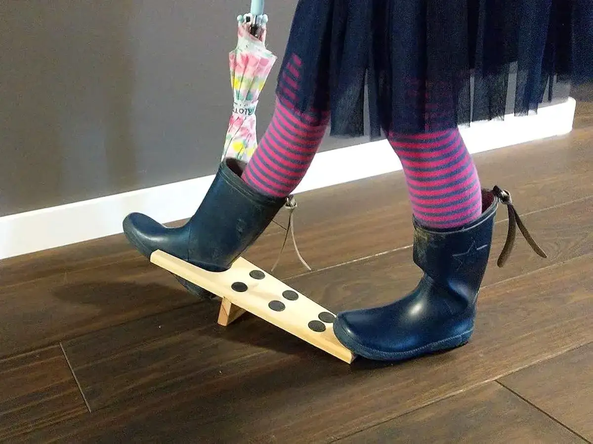 Kind versucht, ihre Gummistiefel anzuziehen. Ein bunter Regenschirm ist im Hintergrund zu sehen.