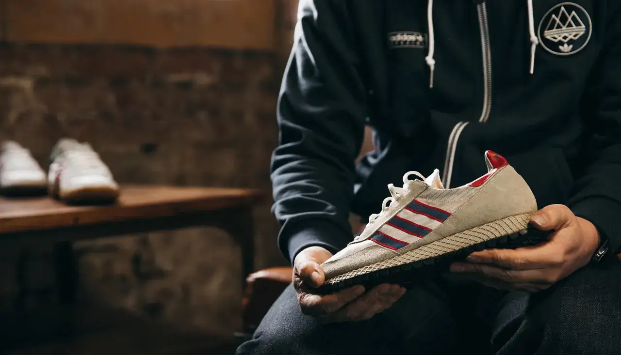 Gary Aspden hält einen beigefarbenen Adidas-Sneaker mit roten und blauen Streifen. Im Hintergrund stehen weitere Sneaker.