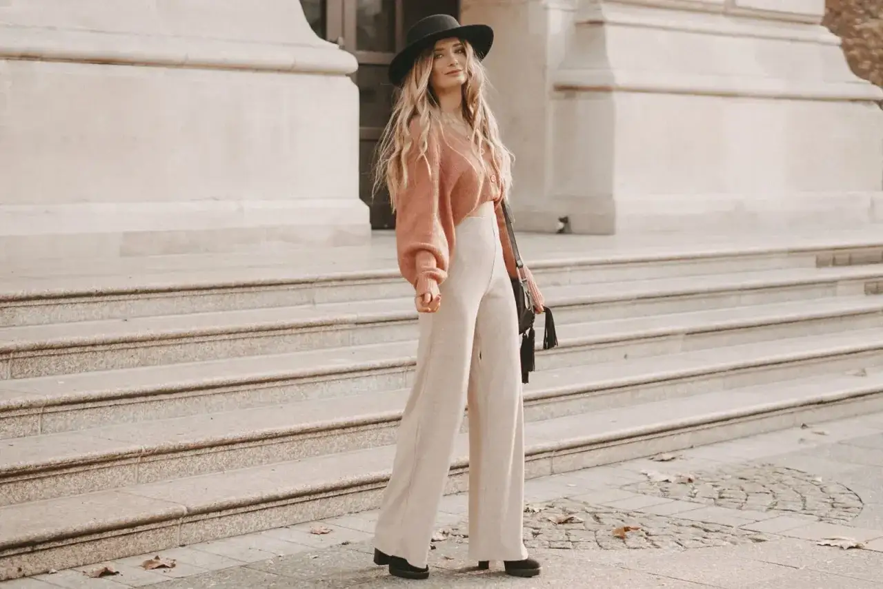 Frau in beigem Strickhose und rosa Cardigan, mit schwarzem Hut und Tasche, steht auf einer Steintreppe. So kann man eine Strickhose kombinieren.