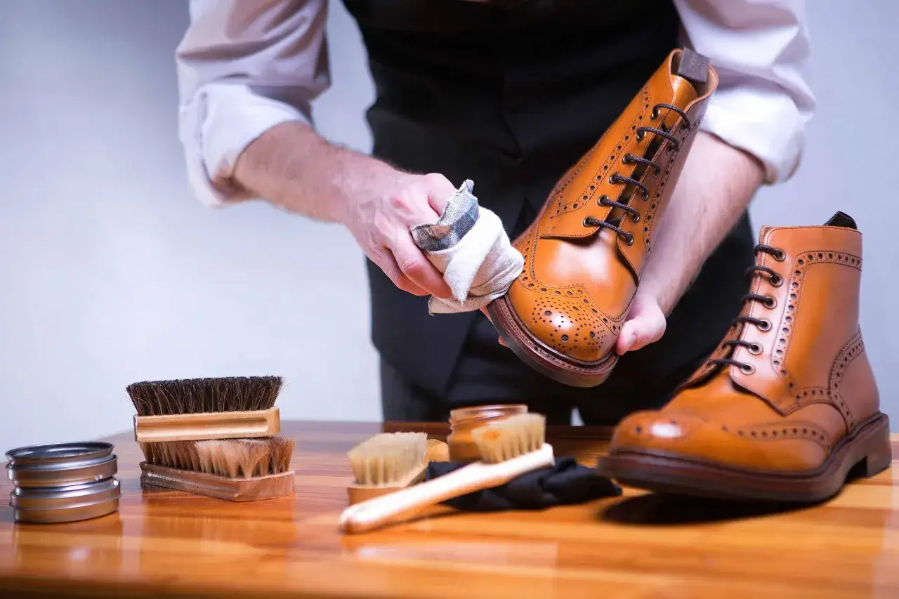 Mann poliert braune Lederschuhe mit einem Tuch. Mehrere B&uuml;rsten und eine Dose Schuhcreme liegen bereit.