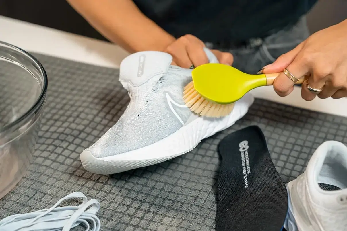 H&auml;nde reinigen mit einer B&uuml;rste die wei&szlig;en Mesh Schuhe. Daneben liegen Schn&uuml;rsenkel und eine Einlegesohle.