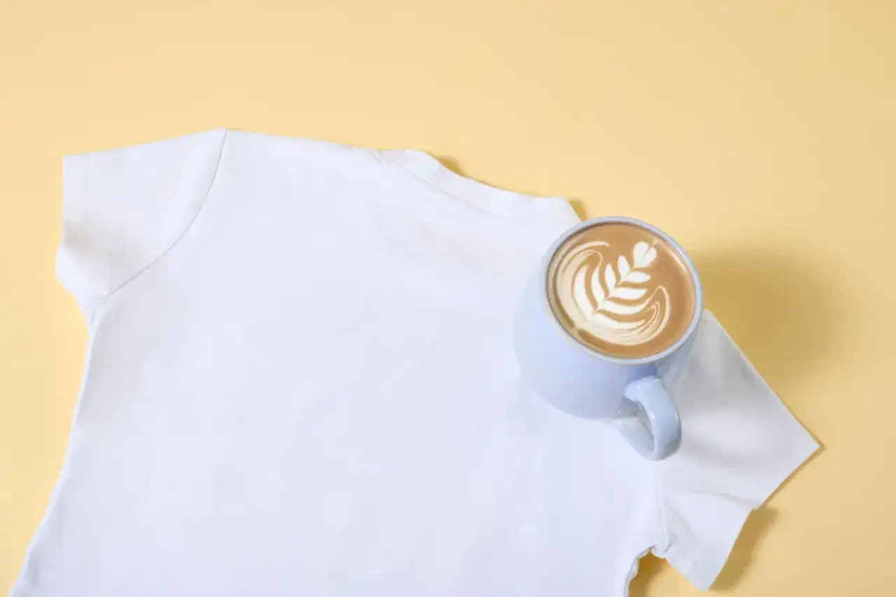 Ein weißes T-Shirt neben einer Tasse Latte Art. Ein kleiner Unfall mit Kaffee kann passieren, aber kaffeeflecken entfernen ist möglich.