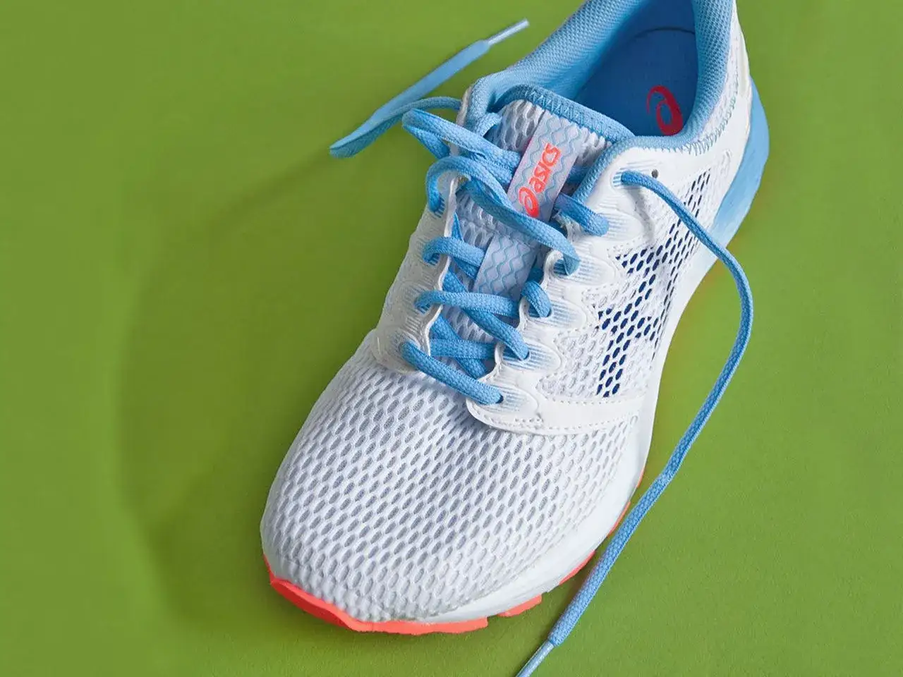 Ein wei&szlig;er Laufschuh mit hellblauen Schn&uuml;rsenkeln, bereit zum Schn&uuml;ren, liegt auf einem leuchtend gr&uuml;nen Hintergrund.