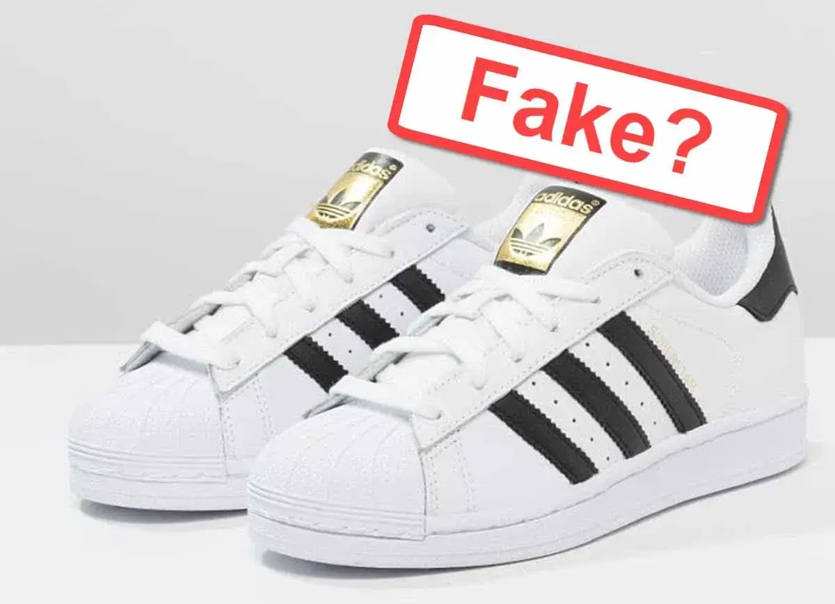 Weiße Adidas Sneaker mit schwarzen Streifen und goldenem Logo. Ein rotes Schild fragt: