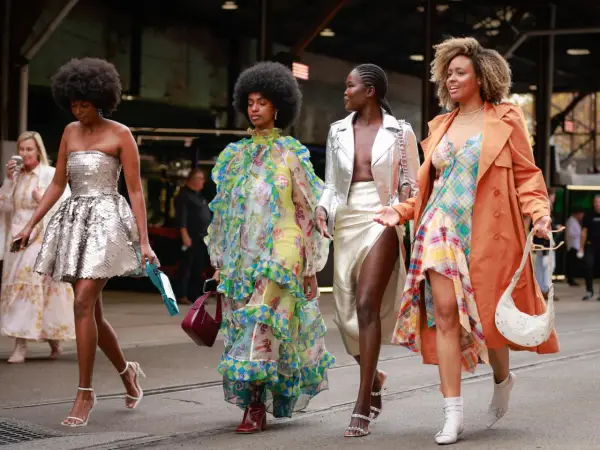 Vier Frauen in auffälligen Outfits. Eine trägt eine silberne Paillettenjacke zum Kleid, eine andere ein buntes Rüschenkleid.