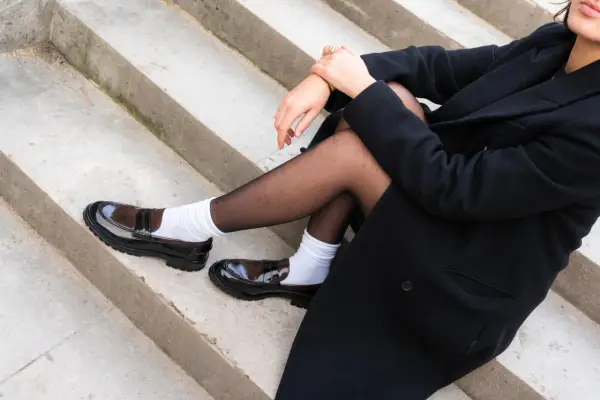 Schwarzer Mantel, schwarze Strumpfhose und weiße Socken mit glänzenden braunen Loafern.